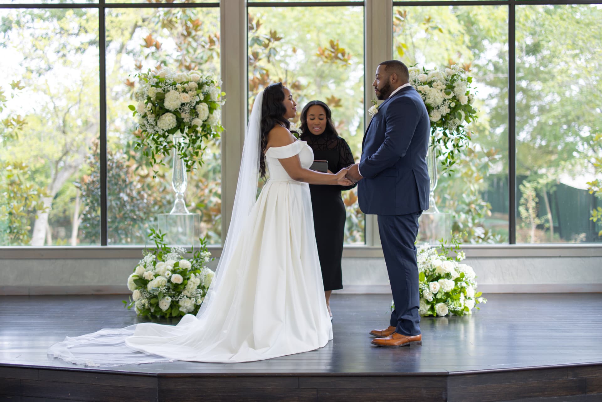 Jennifer Allen officiating a ceremony in front of tall windows and white floral arrangements